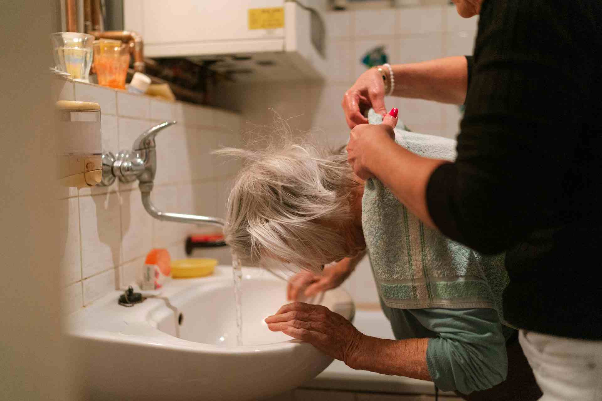 Une femme âgée aux cheveux gris reçoit l'aide d'une soignante dans une salle de bains.